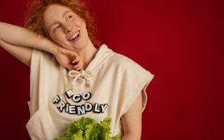 Cheerful woman with red hair in eco-friendly attire holding fresh lettuce against a vibrant red background.