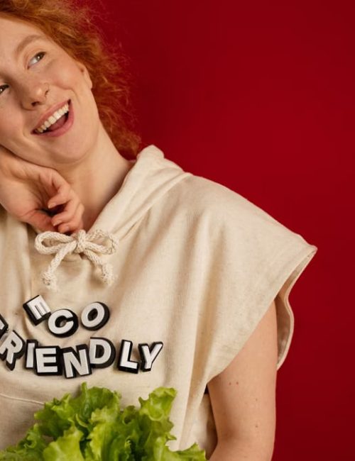 Cheerful woman with red hair in eco-friendly attire holding fresh lettuce against a vibrant red background.