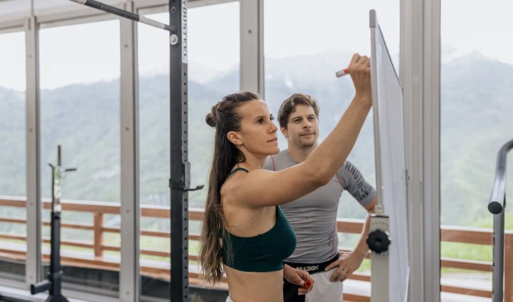 Female fitness coach writes on whiteboard as a male athlete watches in a gym.